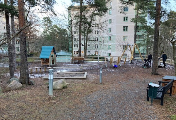 En lekplats med lite barrträd runt om och flerfamiljshus i bakgrudnen.