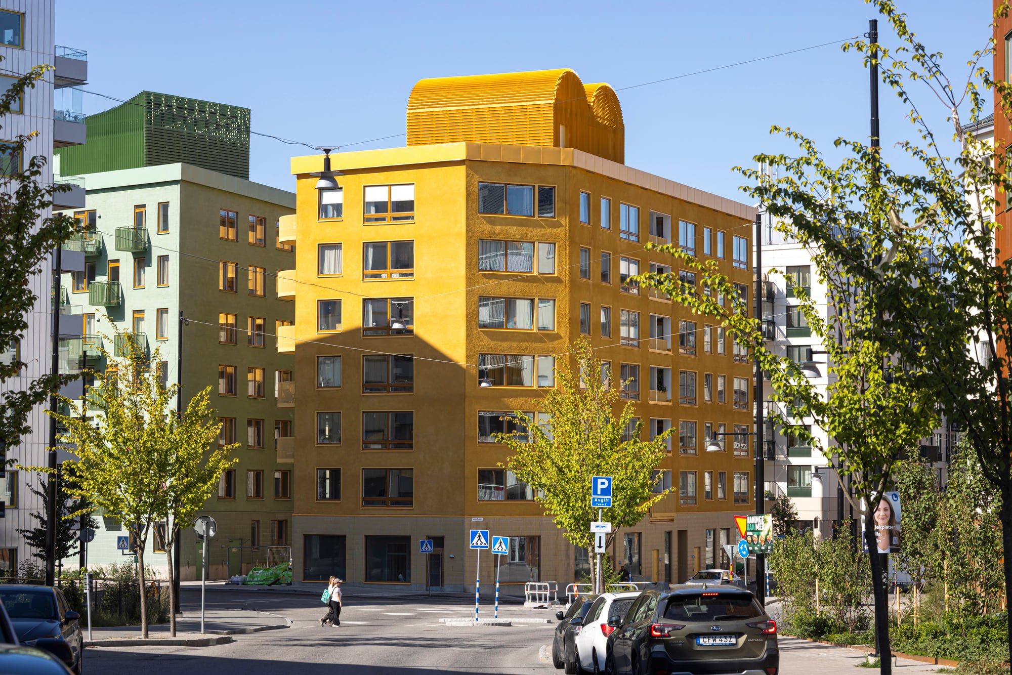Ett gult flerfamiljshus med stora fönster och välvd takkonstruktion, omgivet av andra bostadshus, träd och parkerade bilar vid en solig stadsgata.