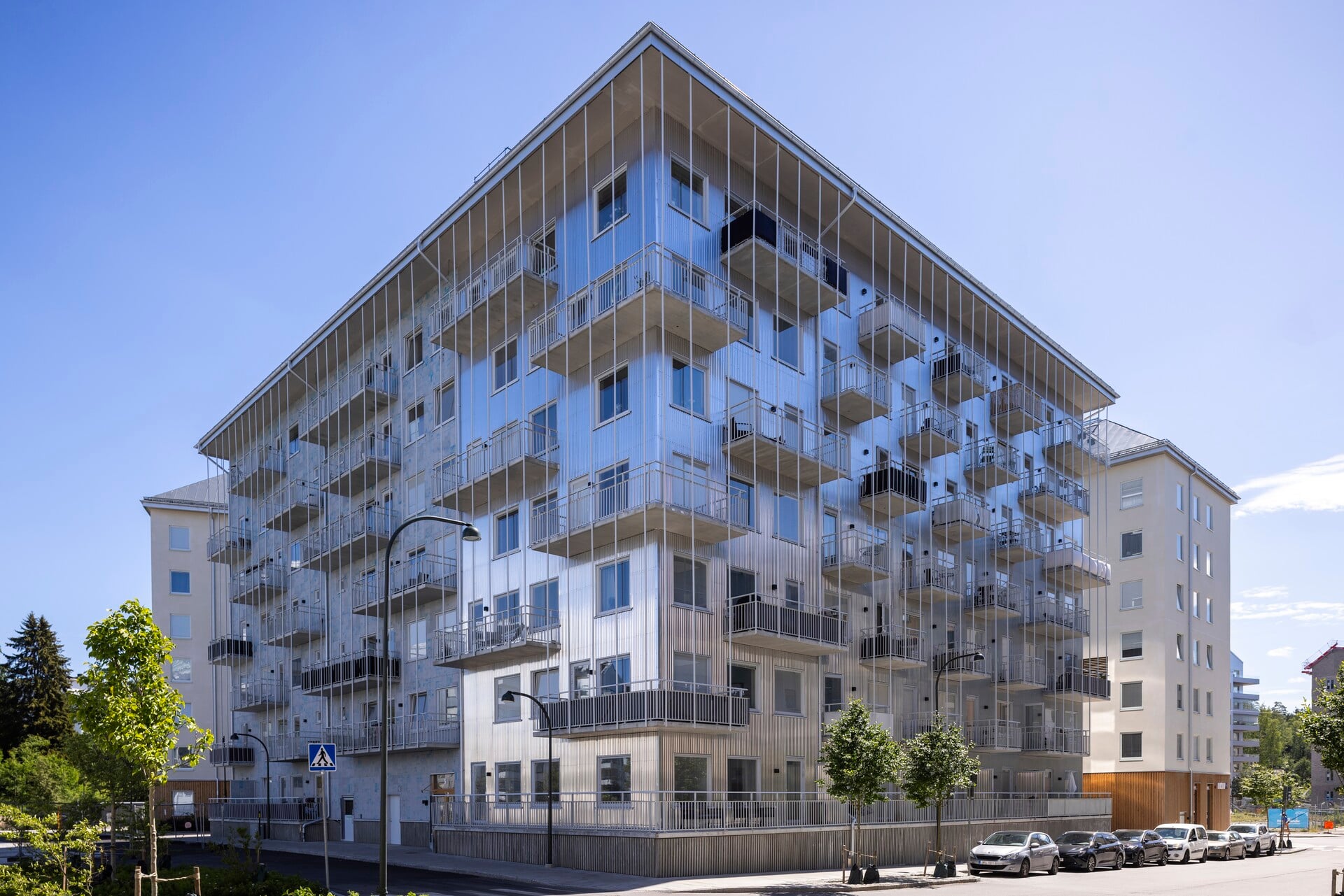 Flerbostadshus i modern stil med fasad i silverfärgad plåt och många utskjutande balkonger, sett från gatan en solig dag med blå himmel, trädplanteringar längs trottoaren och parkerade bilar framför huset. Supernova i Vega Haninge