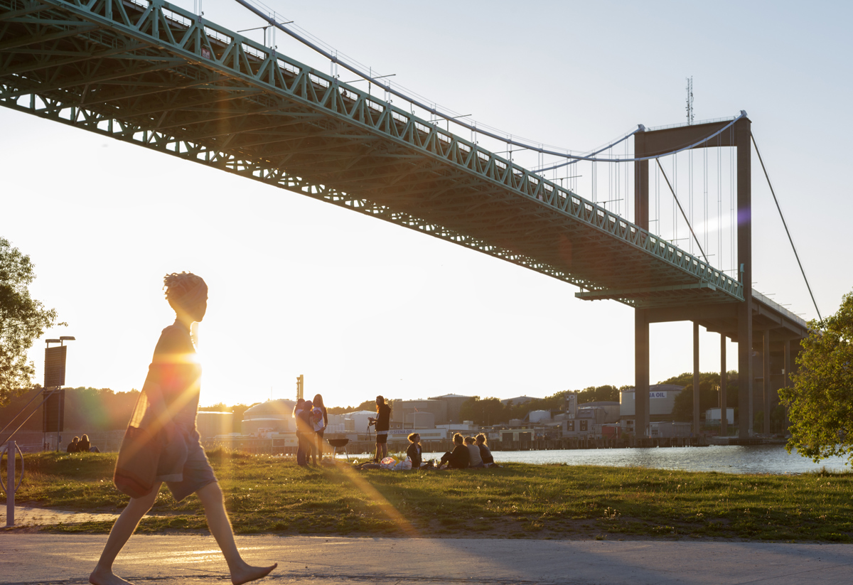 En person går längs en stig vid vattnet med en stor bro i bakgrunden och solen som skiner genom brostrukturen. Flera människor sitter på gräset nära vattnet.