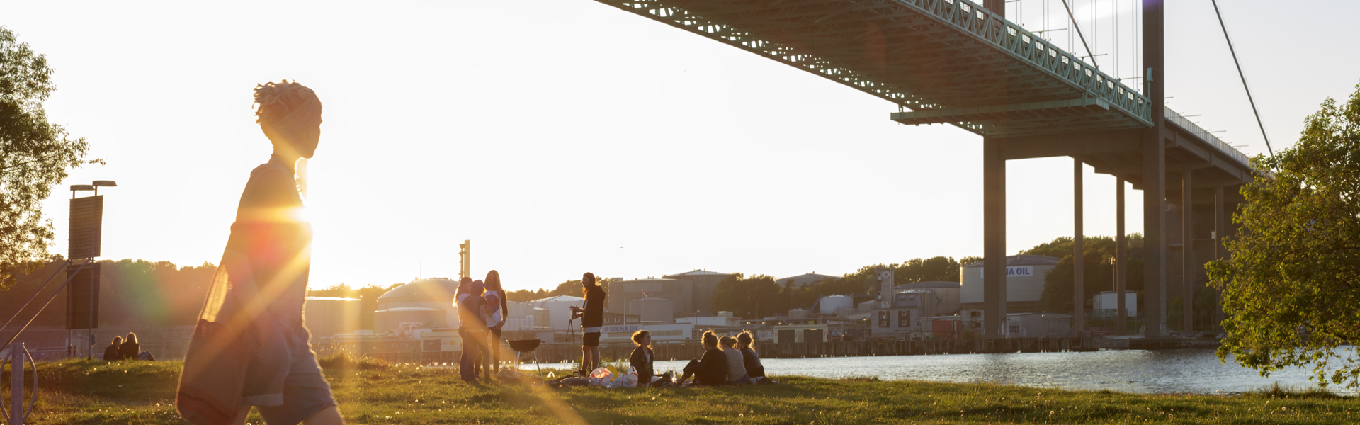 En person går längs en stig vid vattnet med en stor bro i bakgrunden och solen som skiner genom brostrukturen. Flera människor sitter på gräset nära vattnet.