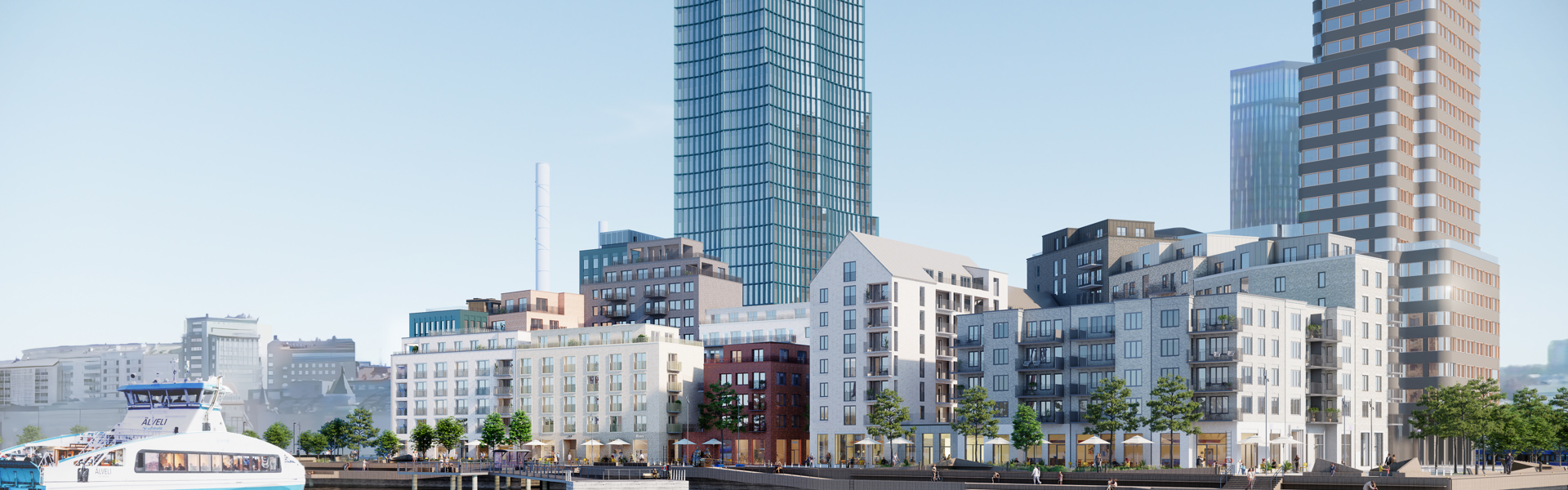 Stadsvy över moderna flerbostadshus och höghus vid vattnet en solig dag. I förgrunden syns en färja på vattnet och kajen kantas av träd och gångvägar. Byggnaderna har varierande höjd och fasader i ljusa färger.