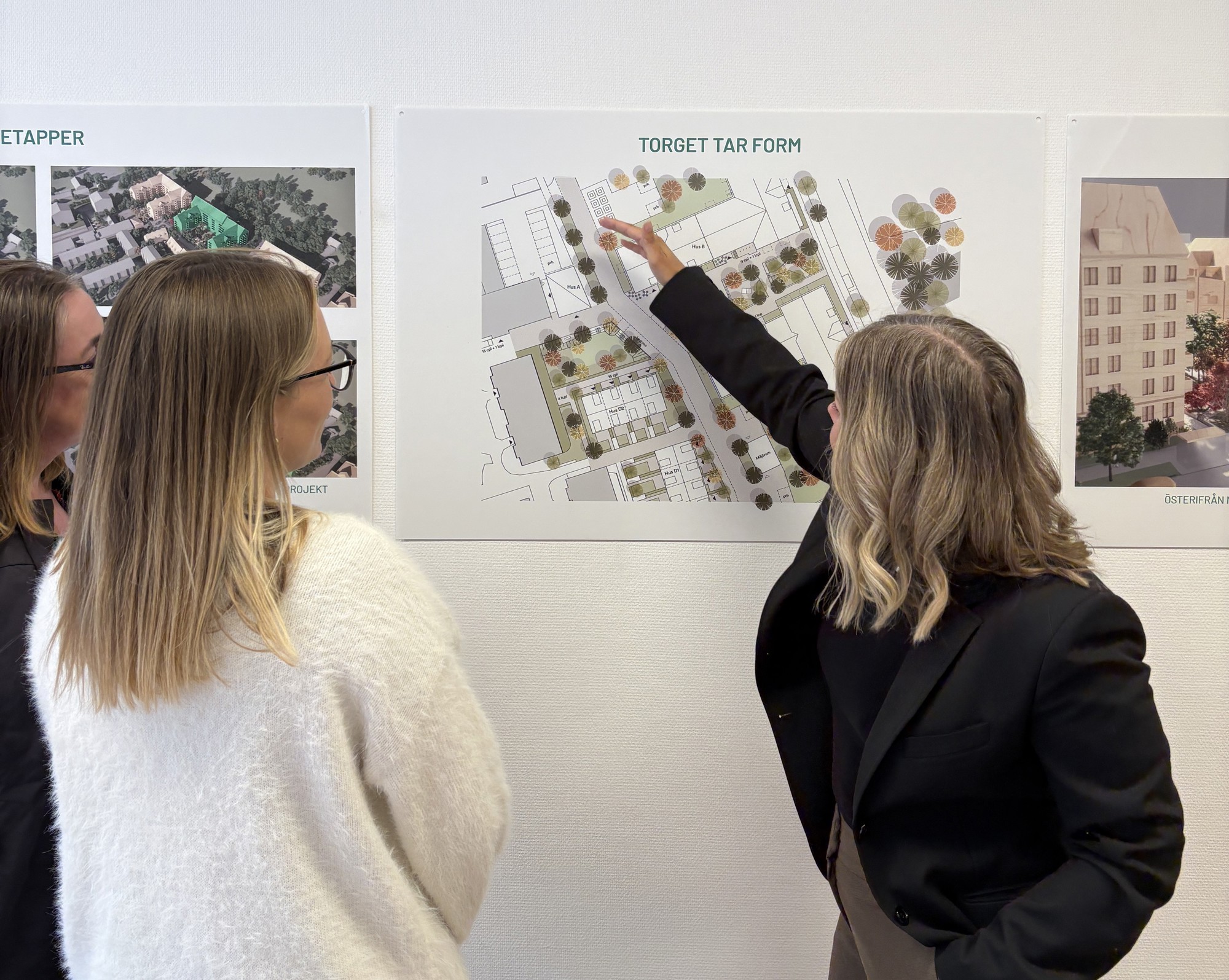 Tre personer står framför en vägg med planscher över ett stadsutvecklingsprojekt. En person pekar på en situationsplan med byggnader, gångstråk och grönområden medan de andra tittar på.
