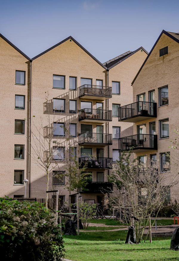 En modern bostadsbyggnad med flera våningar, ljus tegelarkitektur och svarta balkongräcken. Byggnaden omges av en grön innergård med nyplanterade träd och buskar. Himlen är delvis molnig, och atmosfären är lugn.