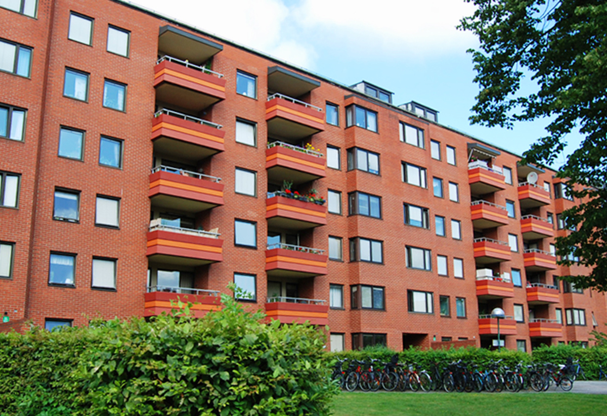 Ett flerbostadshus i rött tegel med flera våningar och balkonger, omgivet av gröna buskar och träd, samt en rad parkerade cyklar i förgrunden.
