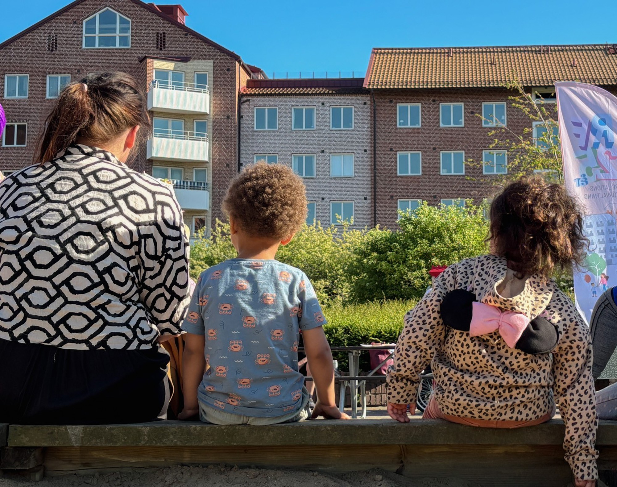 Tre personer sitter på en bänk med ryggen mot kameran och tittar mot flerbostadshus och grönska i soligt väder. Bilden visar en vuxen och två barn på en innergård med bostadshus i bakgrunden.