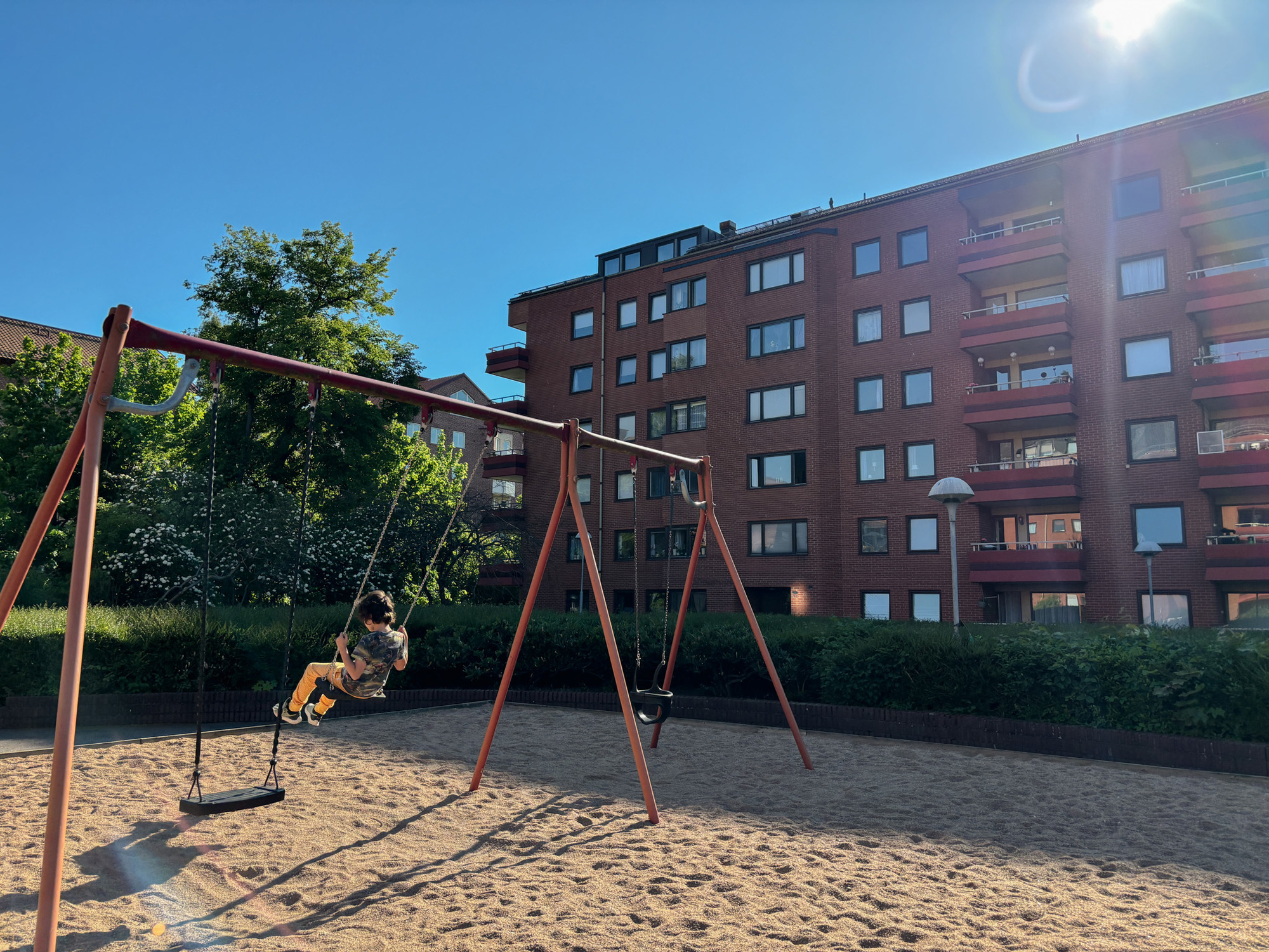 Ett barn som gungar på en gungställning i en sandlåda framför ett rött flerbostadshus en solig dag, omgiven av gröna buskar och träd.