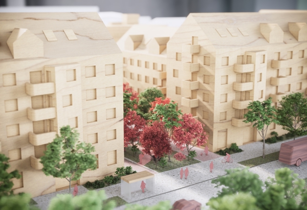 En närbild på en arkitekturmodell som visar flera flerbostadshus i trä, omgivna av träd och grönska. Mellan husen finns en gångväg med röda och gröna träd samt små figurer som föreställer människor. Modellens detaljer illustrerar en trivsam och grön stadsmiljö med fokus på boende och utemiljö.