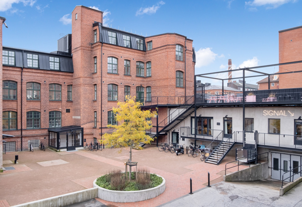 Teglad industribyggnad i flera våningar med stora fönster och svart tak, ombyggd till modern kontors- och mötesmiljö. I förgrunden en innergård med träd i planteringsrondell, cyklar parkerade längs fasaden och en trappa upp till en terrass där skylten ”SIGNAL” syns på väggen.