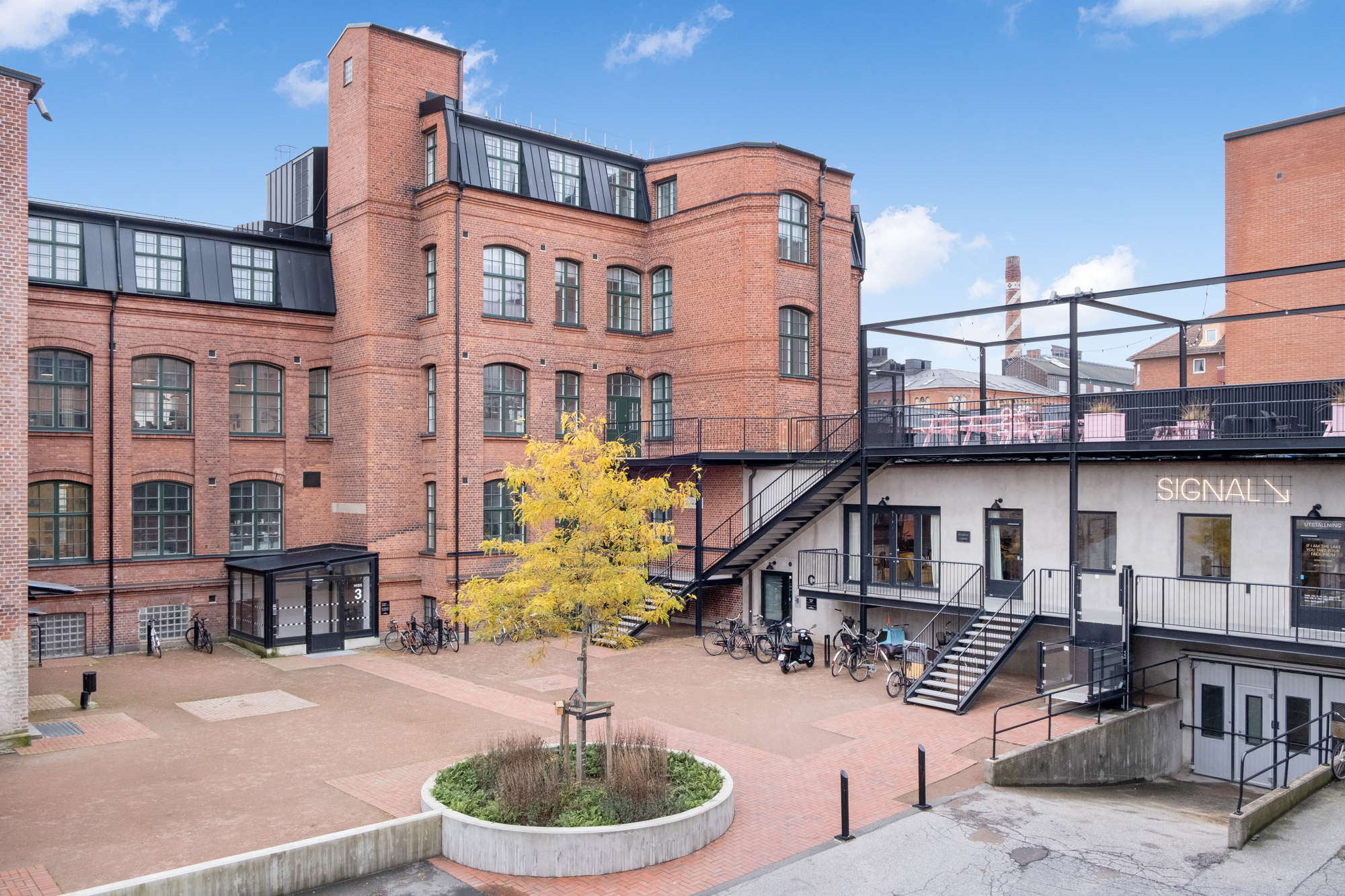 Teglad industribyggnad i flera våningar med stora fönster och svart tak, ombyggd till modern kontors- och mötesmiljö. I förgrunden en innergård med träd i planteringsrondell, cyklar parkerade längs fasaden och en trappa upp till en terrass där skylten ”SIGNAL” syns på väggen.