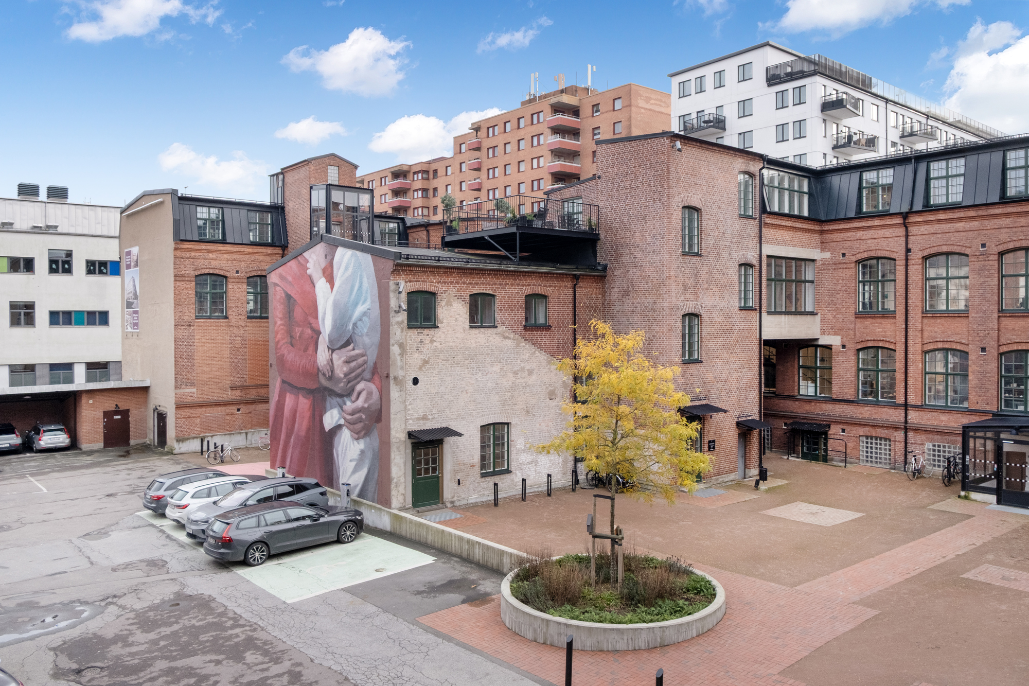  Utegård vid en äldre tegelbyggnad med stora fönster, där en fasad pryds av en färgstark muralmålning av två omfamnande personer. I förgrunden syns en liten planterad rondell med ett träd, parkerade bilar och en asfalterad gård, med högre bostadshus i bakgrunden mot en blå himmel med moln.