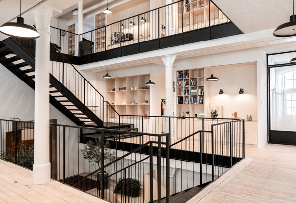 Ljust och modernt kontorsutrymme i flera våningar med öppna ytor, svarta räcken och en bred trappa i metall. Väggarna är vita med inbyggda bokhyllor och hängande svarta lampor. Golvet är i ljust trä, och stora fönster släpper in naturligt ljus.
