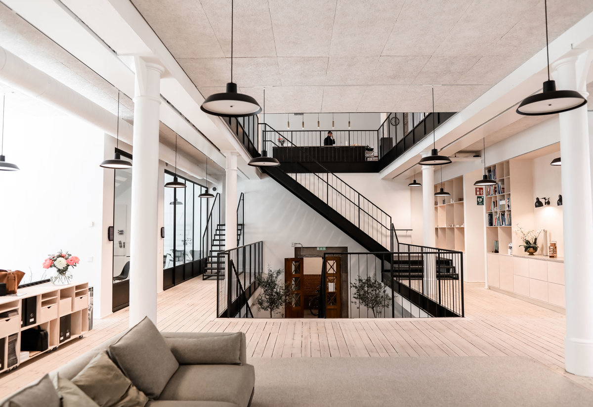 Modernt, öppet kontorslandskap med ljusa väggar, stora fönster och svarta trappor i metall som leder till ett övre våningsplan. I förgrunden syns en beige soffa och inredning med förvaringsmöbler och blommor. Rummet är upplyst av flera hängande svarta taklampor.