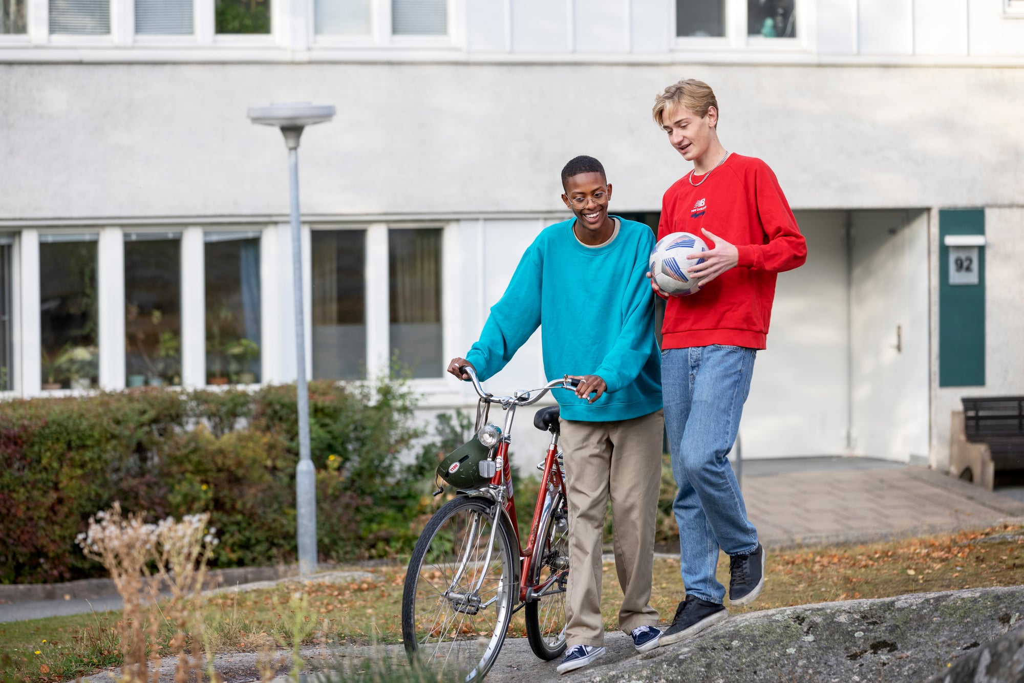 Två personer går tillsammans utomhus framför ett bostadshus. Den ena leder en cykel och den andra håller i en boll.