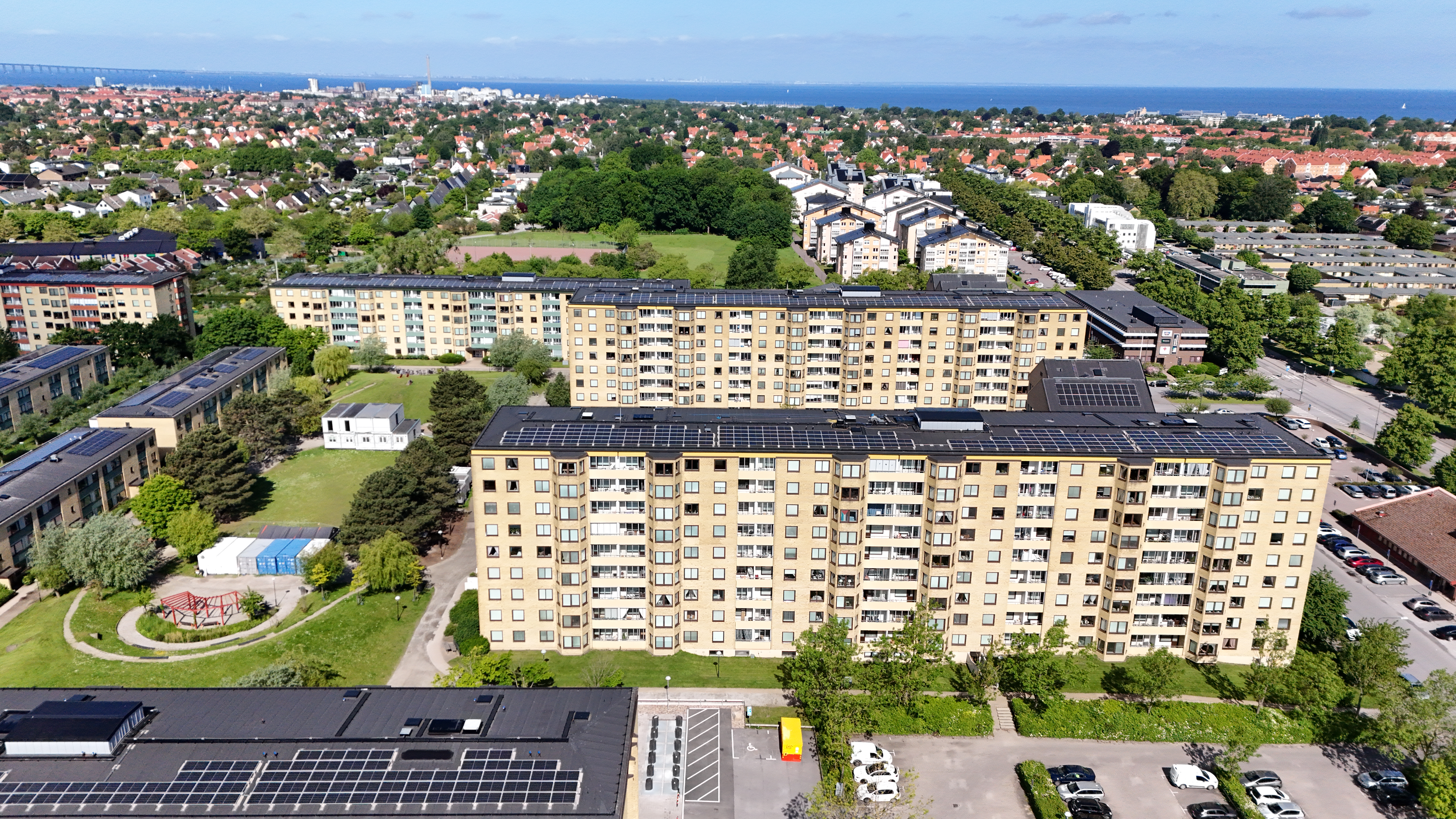 Ett flerbostadshus i gult tegel sett uppifrån i ett bostadsområde med grönområden och fler liknande hus runtom; en röd markering visar en specifik lägenhet på mitten av den långa fasaden.