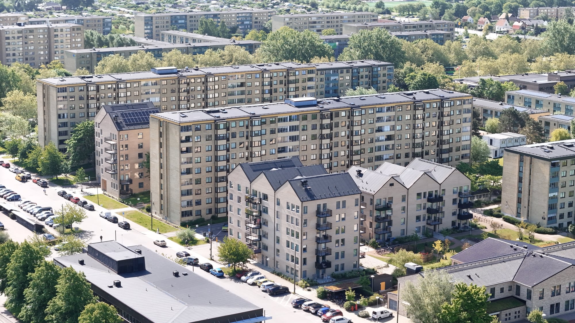 Flygbild över ett bostadsområde med flera flerbostadshus, gator, parkerade bilar och grönska. I bakgrunden syns fler byggnader och träd under en ljus himmel.