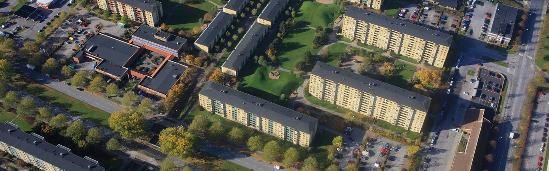 Flygbild över ett bostadsområde med flera flerfamiljshus och grönområden. Husen är omgivna av träd och parkeringsplatser, och det finns gångvägar som förbinder byggnaderna.