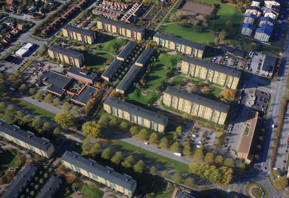 Flygbild över ett bostadsområde med flera flerfamiljshus och grönområden. Husen är omgivna av träd och parkeringsplatser, och det finns gångvägar som förbinder byggnaderna.