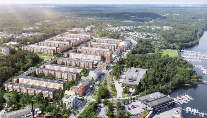 Flygfoto över ett bostadsområde omgivet av skog, med flerfamiljshus i rader, marina med båtar vid vattnet till höger och vägar som slingrar sig genom landskapet.