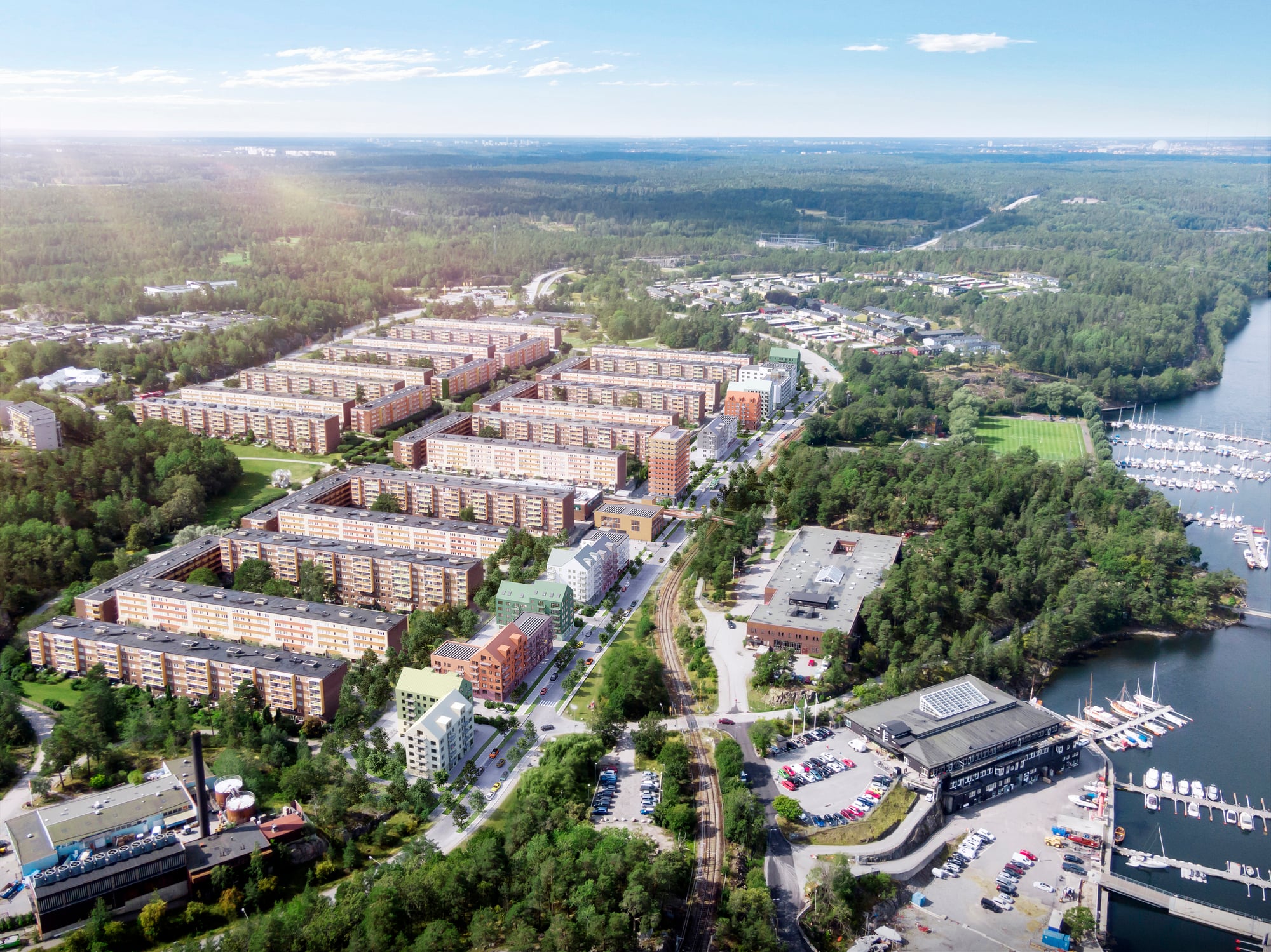 Flygfoto över ett bostadsområde omgivet av skog, med flerfamiljshus i rader, marina med båtar vid vattnet till höger och vägar som slingrar sig genom landskapet.