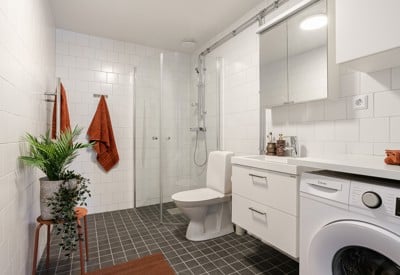 Vit, modern badrumsmiljö med duschhörna i glas, toalett, vit kommod med spegelskåp, tvättmaskin och mörkgrå klinkergolv samt rostfärgade handdukar som accentfärg.