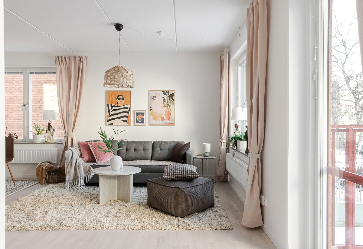 Ljust vardagsrum med grå soffa, brun sittpuff och runt soffbord på ljus ryamatta, stora fönster med beige gardiner samt tavlor och gröna växter som dekoration.
