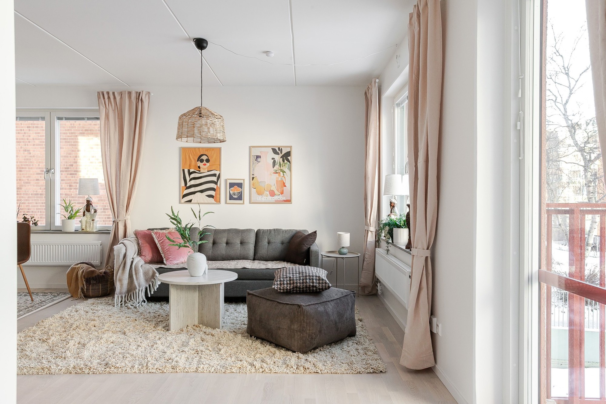 Ljust vardagsrum med grå soffa, brun sittpuff och runt soffbord på ljus ryamatta, stora fönster med beige gardiner samt tavlor och gröna växter som dekoration.