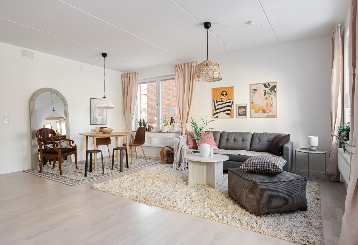 Ljust vardagsrum med öppen planlösning mot matplats, grå soffa och brun sittpuff på ljus ryamatta, matbord med stolar, stora fönster med beige gardiner samt tavlor och växter som dekoration.