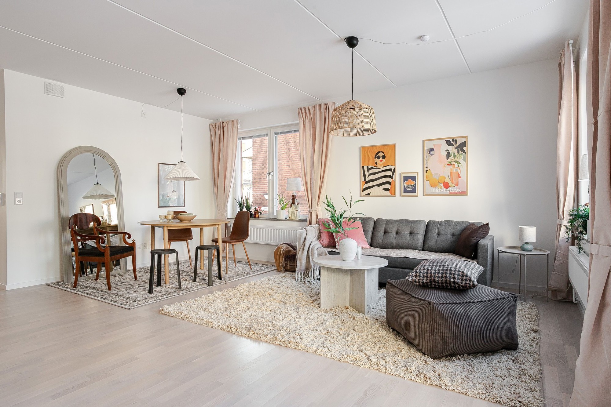Ljust vardagsrum med öppen planlösning mot matplats, grå soffa och brun sittpuff på ljus ryamatta, matbord med stolar, stora fönster med beige gardiner samt tavlor och växter som dekoration.