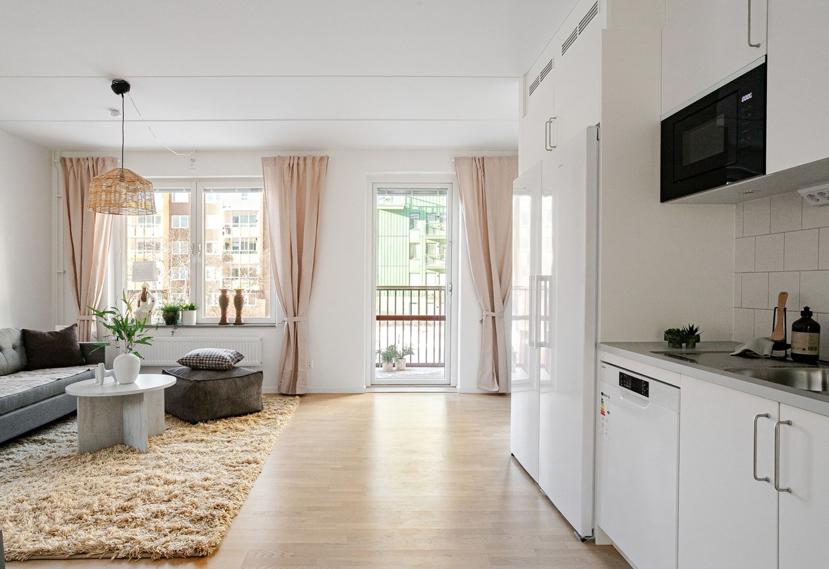 Ljust vardagsrum med öppen planlösning mot vit köksdel, grå soffa och soffbord på ljus ryamatta samt stora fönster och balkongdörr med beige gardiner.