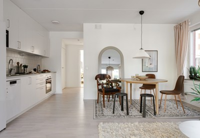 Ljust kök med vita skåp och bänkskiva till vänster samt matplats med runt bord, blandade stolar och taklampa på en matta framför en spegel.