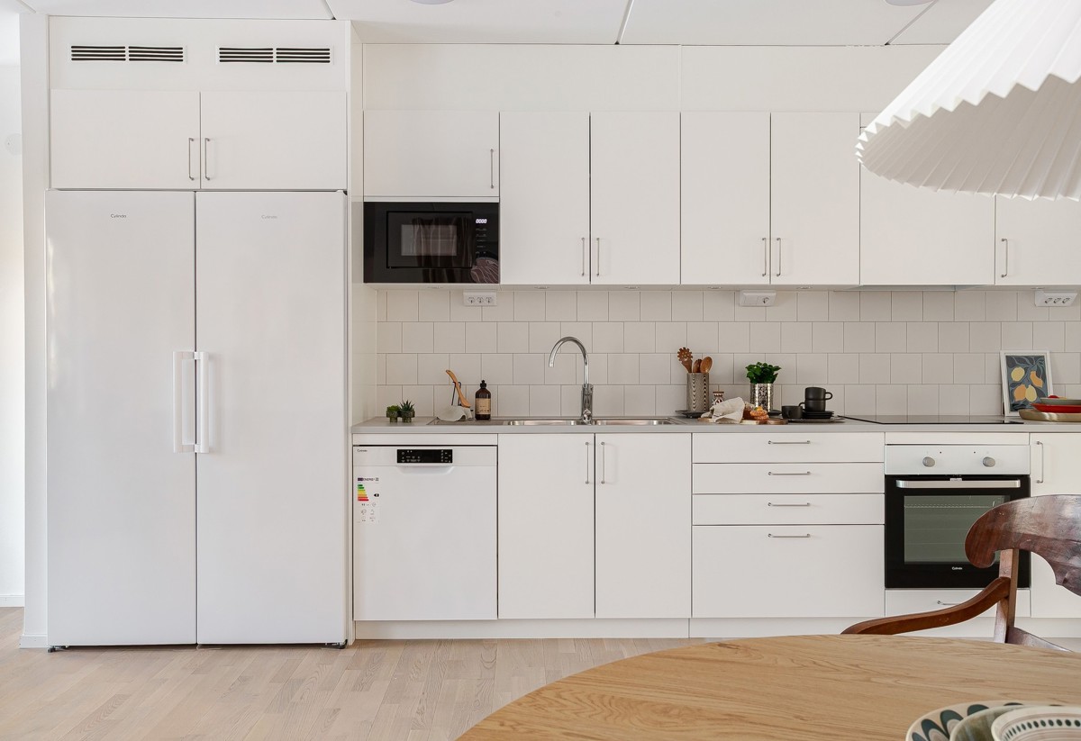 Ljust, modernt kök med vita skåp i rad, vit kyl och frys, inbyggd mikro, diskmaskin och ugn samt ljus bänkskiva med köksredskap och växt som dekoration.