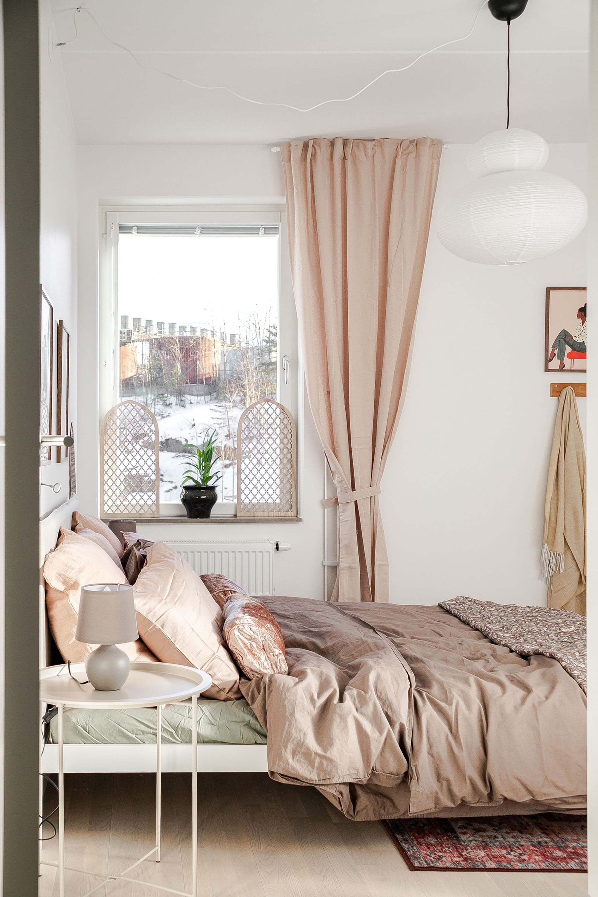 Ljust sovrum med bäddad säng i beige textilier, nattduksbord med lampa, stort fönster med beige gardin och dekor med växter i fönstret.