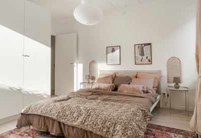 Ljust sovrum med bäddad säng i beige och bruna textilier, två nattduksbord med lampor, vita garderober till vänster och tavlor samt dekorativa väggpaneler ovanför sänggaveln.