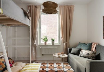 Mysigt mindre rum med loftsäng, grå bäddsoffa, små soffbord, fönster med beige gardiner och gröna växter i fönstret samt mönstrad matta på golvet.