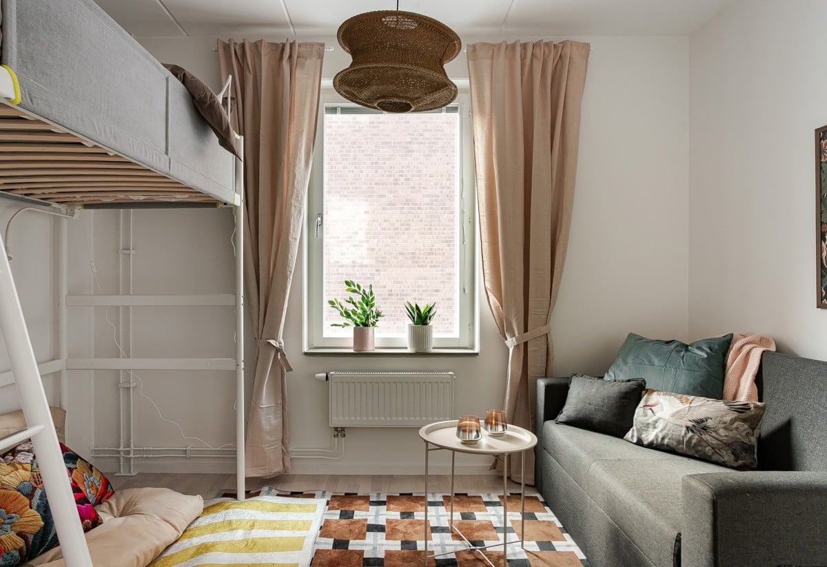 Mysigt mindre rum med loftsäng, grå bäddsoffa, små soffbord, fönster med beige gardiner och gröna växter i fönstret samt mönstrad matta på golvet.