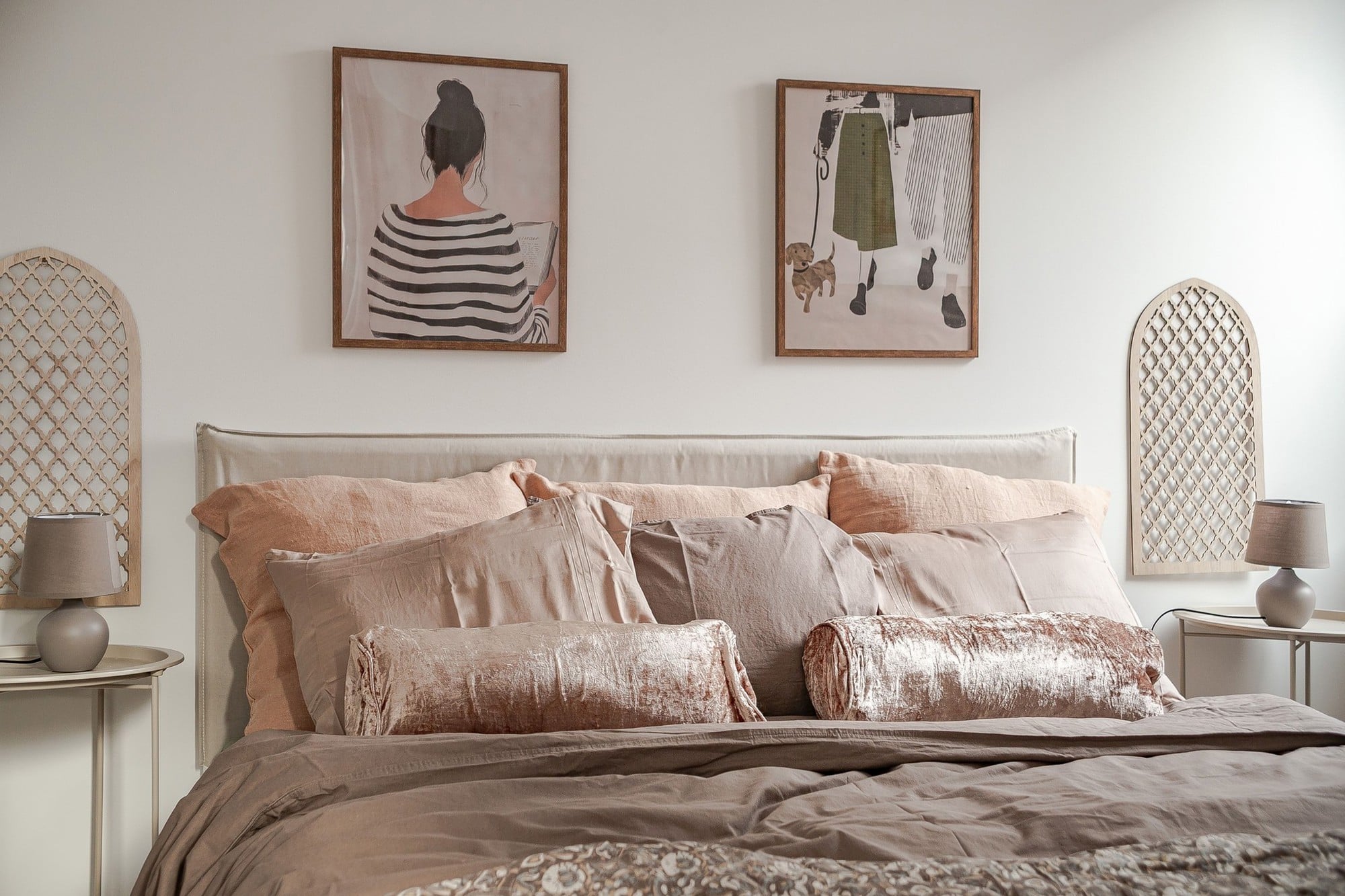 Närbild på bäddad dubbelsäng i beige och rosatonade textilier med dekorationskuddar, två små nattduksbord med lampor samt två tavlor och dekorativa väggpaneler ovanför sänggaveln.