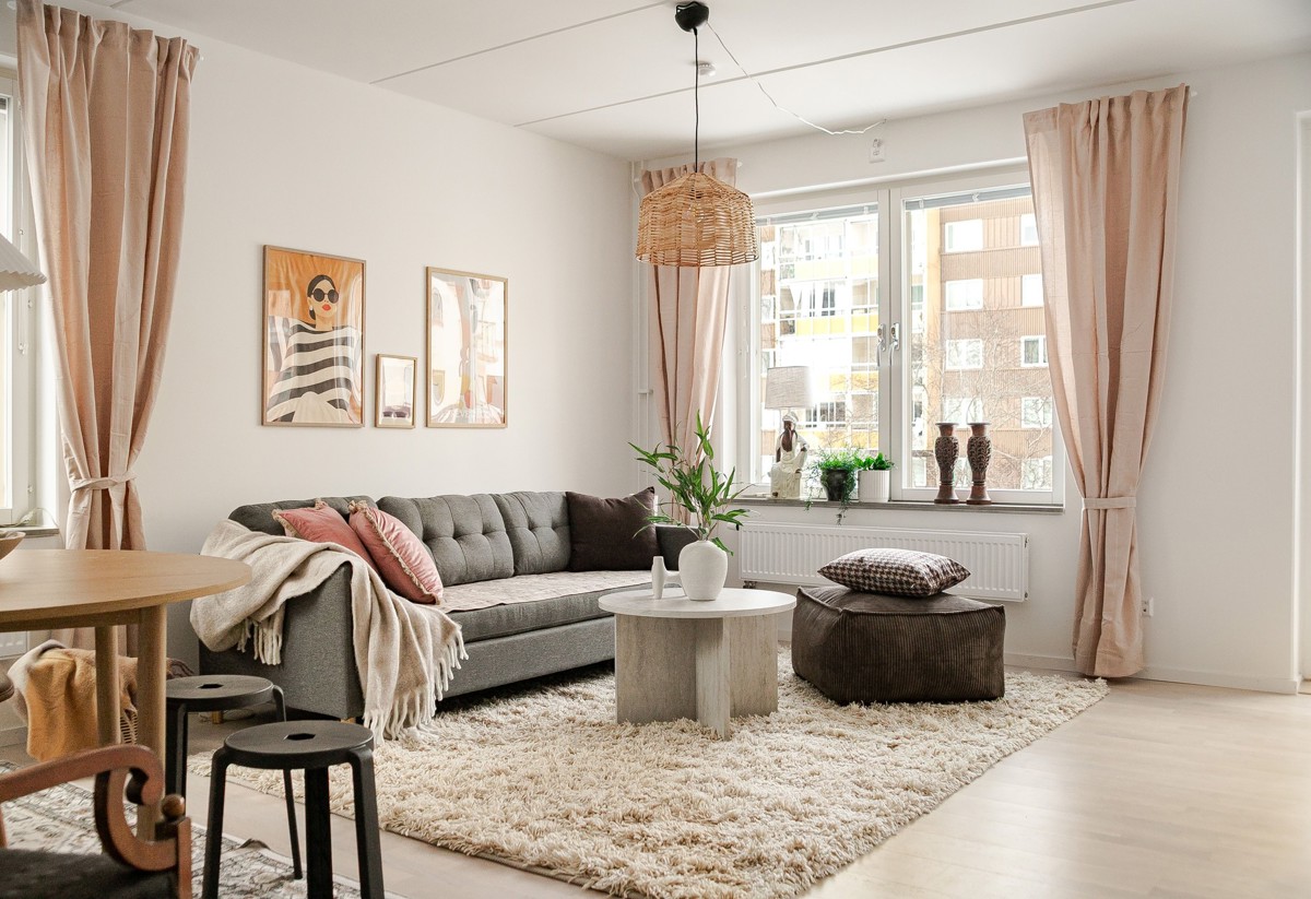 Ljust vardagsrum med grå soffa, runt soffbord och brun sittpuff på ljus ryamatta, stora fönster med beige gardiner samt tavlor och gröna växter som dekoration.
