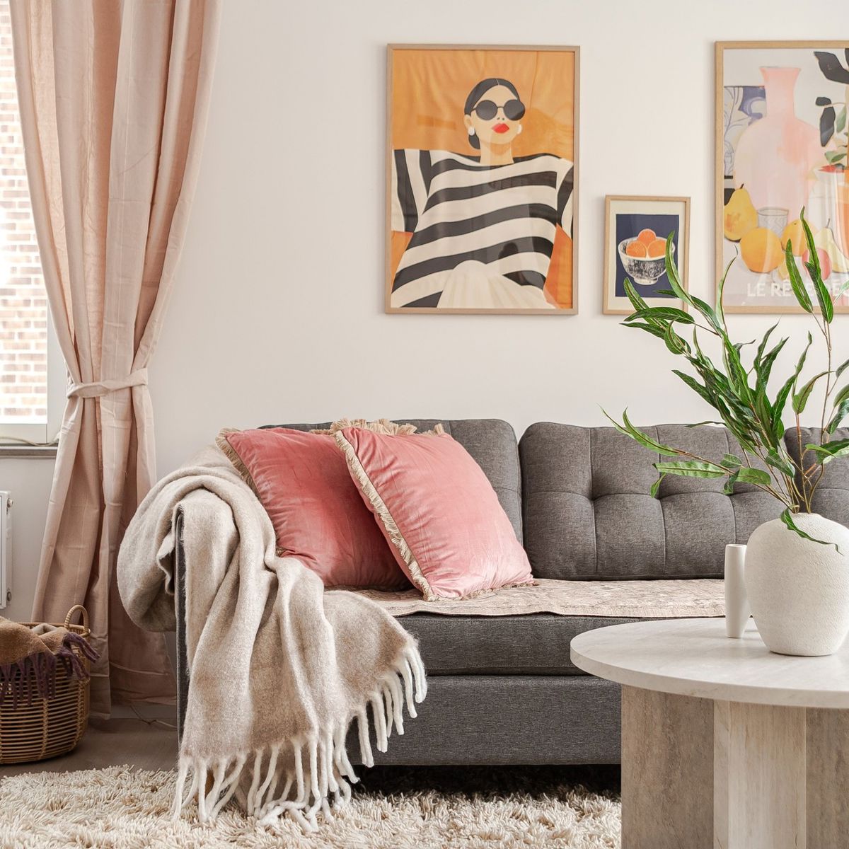 Ljust vardagsrum med grå soffa, rosa prydnadskuddar och pläd, runt soffbord med vit vas och grön växt, samt fönster med beige gardiner och färgstarka tavlor på väggen.