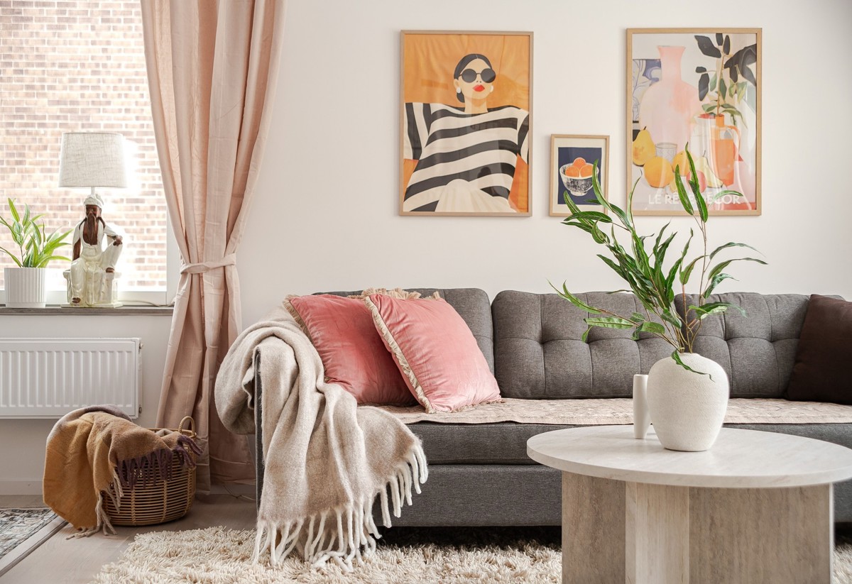Ljust vardagsrum med grå soffa, rosa prydnadskuddar och pläd, runt soffbord med vit vas och grön växt, samt fönster med beige gardiner och färgstarka tavlor på väggen.