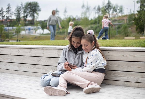 Två tjejer som tittar på telefonen i Vega med en familj som spelar fotboll i bakgrunden