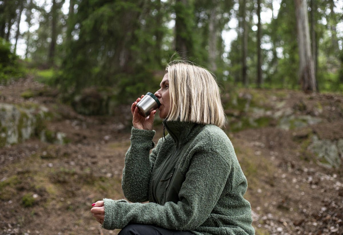 Kvinna i grön jacka dricker kaffe ur termoskopp i skogen